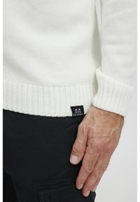 Jersey de punto blanco con un dobladillo y puños acanalados. Presenta un patrón texturizado y una etiqueta negra con texto blanco en el lateral.