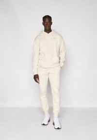 Hoodie couleur crème avec une poche avant, associé à un jogging assorti. Présente un tissu texturé et des accents de logo subtils. Porté avec des baskets blanches.