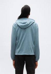 Άτομο που φαίνεται από πίσω φοράει ανοιχτό μπλε μπουφάν με κουκούλα και μαύρο παντελόνι σε απλό λευκό φόντο.