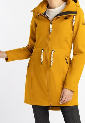 Femme portant un imperméable jaune moutarde zippé avec cordons blancs et capuche, associé à un pantalon foncé.