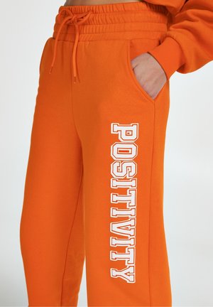 Pantalones deportivos naranjas hechos de tela suave, con un ajuste cómodo, cintura elástica y bolsillos laterales. Texto "POSITIVITY" impreso en blanco con alineación vertical.