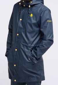 Marinblå vattentät jacka med huva, guldknappar, justerbara dragsnören och sidofickor. Slät textur med en logotyp på bröstet.