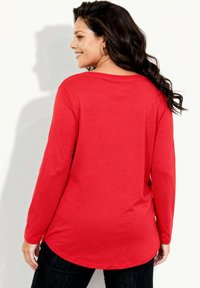 Czerwona koszula z długim rękawem o zaokrąglonym dole, wykonana z miękkiego materiału. Posiada dekolt z tyłu i jest zestawiona z ciemnymi dżinsami.