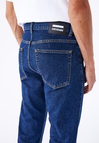 Jeans de denim azul oscuro con corte recto, costuras visibles y una etiqueta negra con "DRDENIM" en la cinturilla trasera.