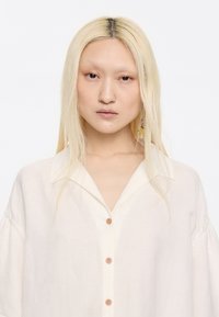 Light cream button-up shirt with a collar, short puffed sleeves, and wooden buttons. Smooth texture and relaxed fit.