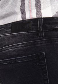 Jean en denim noir avec un patch logo en cuir à la taille. Présente des coutures discrètes et une texture lisse.