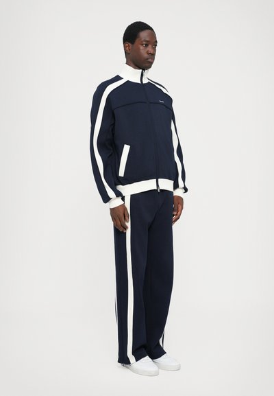 Training suit bleumarin cu dungi albe pe jachetă și pantaloni; jachetă cu fermoar și guler înalt, cu buzunare laterale; material din amestec de bumbac.
