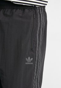 Melni sporta bikses ar elastīgu jostasvietu, rāvējslēdzēja kabatu un trim vertikālām svītrām sānos. Funkcijā Adidas logo pelēkā krāsā.