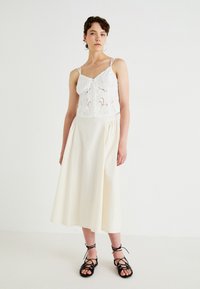 Hvit, brodert crop top med tynne stropper, sammen med en flytende, kremfarget midi-skjørt. Sorte snøresandaler fullfører antrekket.