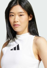 Vit ärmlös topp med hög krage och svart logotypgrafik på bröstet. Slät tyggtextur och figurnära design, med en röd tatuering på axeln.