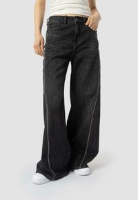 Szerokie czarne dżinsy wykonane z denimu, z białymi szwami wzdłuż szwów bocznych i kieszeni. Model nosi białe sneakersy.