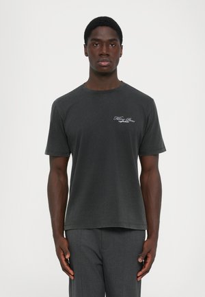 Man wearing a faded black short-sleeve t-shirt with white text on the chest, paired with dark gray trousers, standing facing forward.