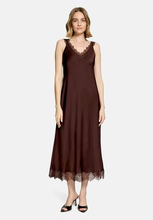 Frau mit schulterlangem Haar, die ein ärmelloses dunkelbraunes Kleid mit Spitzenbesatz und schwarze offene Peep-Toe-Absätze trägt, steht vor einem schlichten Hintergrund.