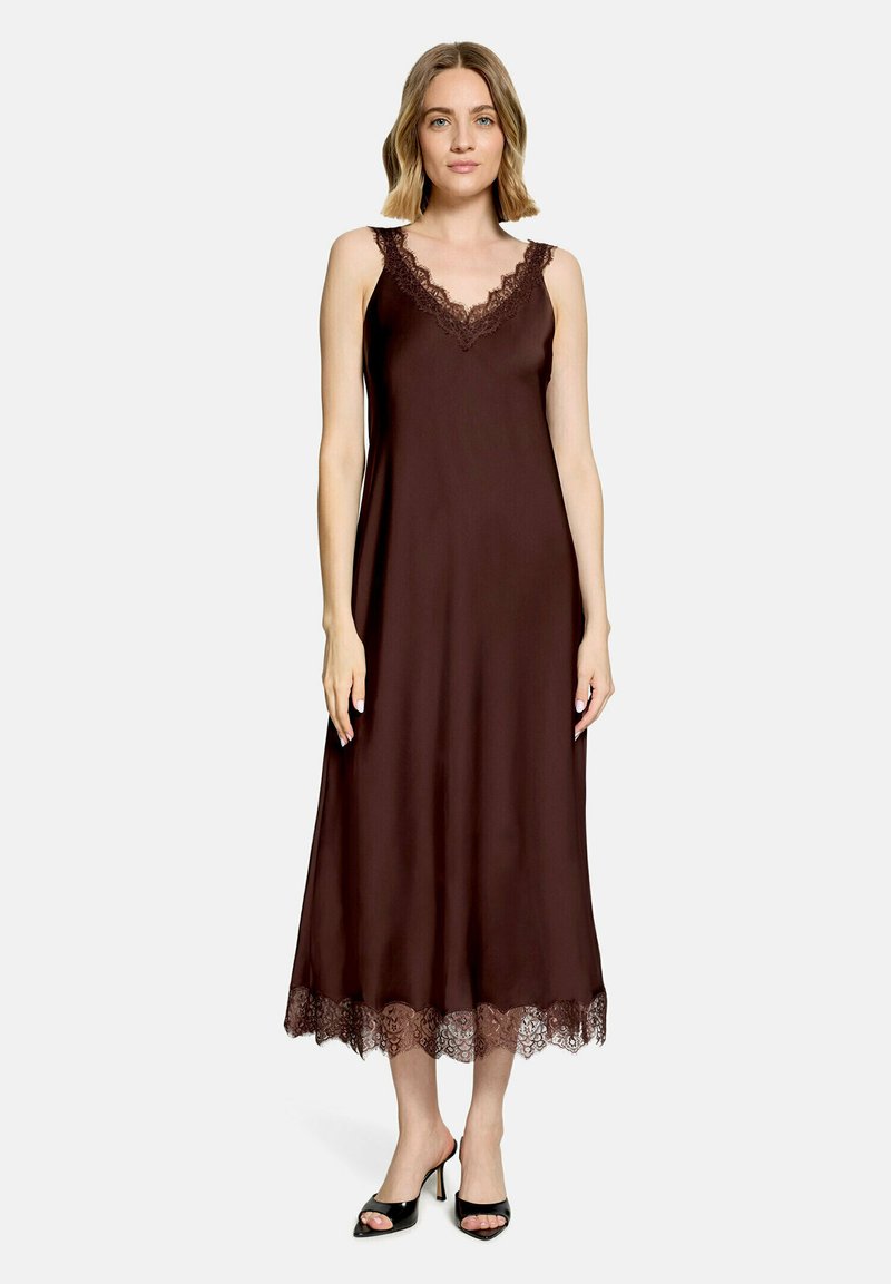 Femme aux cheveux mi-longs portant une robe sans manches marron foncé avec bordure en dentelle et des talons ouverts noirs, debout devant un fond uni.