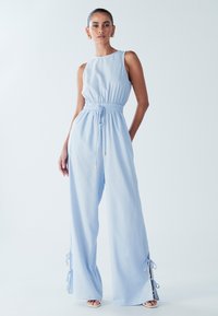 Svetlo modra kombineža z oprijetim zgornjim delom in elastičnim pasom. Ima široke hlače s stranskimi razporki in vrvicami za zavezovanje. Iz mehke, tekoče tkanine.