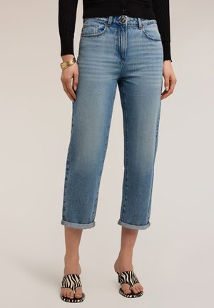 Vrouw met lichtblauwe omgeslagen cropped jeans, zwarte top met lange mouwen, sandaal met hoge hakken in zebraprint en gouden armbanden aan haar linkerpols.