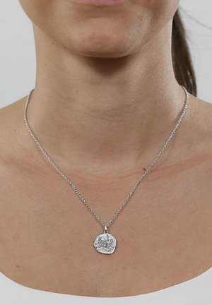 Nahaufnahme des Halses einer Person, die eine silberne Halskette mit einem runden, strukturierten Anhänger trägt, der mit einem Blumendesign graviert ist.