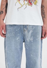 Camiseta blanca con gráfico de dragón, combinada con pantalones de mezclilla azul claro que presentan diseños tonales blancos y un corte suelto.