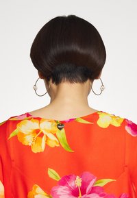 Korte mouwen bloes in helder oranje met roze en gele bloemenpatronen. Heeft een hartvormig label aan de halslijn en ronde oorbellen.