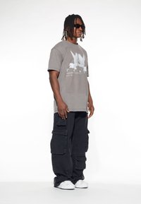 Grå t-shirt i bomull med vit duv-grafik och text, matchad med svarta cargobyxor med flera fickor och sidoflappar. Vita sneakers.