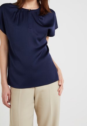 Vrouw draagt een navyblauwe blouse met korte mouwen, een ronde hals met knopen en beige hoge taille broek.