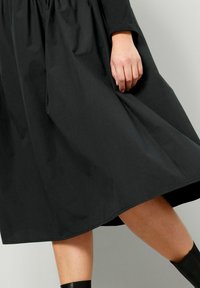 Černé šaty do kolen vyrobené z jemné tkaniny s rozšířenou sukní a dlouhými rukávy. Spodní lem je rovný a ukazuje hladkou strukturu.