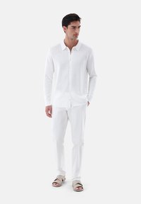 Weißes Langarmhemd mit strukturiertem Muster, geknöpfter Front, kombiniert mit weißen Hosen und hellen Sandalen.