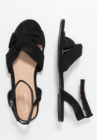 Sandales en daim noir avec un design à l'avant noué, bout ouvert et bride de cheville réglable. Semelle plate et doublure intérieure beige lisse.