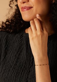 Gouden armband met verspreide zwarte kralen, om een pols. De achtergrond heeft een gestructureerde zwarte stof met een gekreukeld ontwerp.