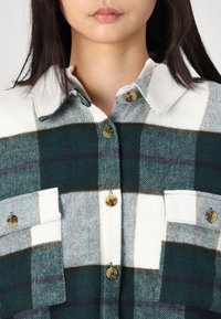 Zielono-biała koszula w kratę z kołnierzykiem, dwoma kieszeniami na piersi i brązowymi guzikami. Tkanina wydaje się miękka o szczotkowanej fakturze.