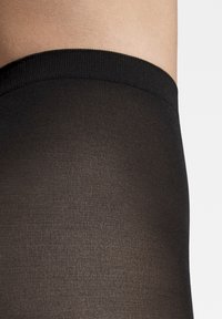 Collants opaques noirs avec une texture lisse et une ceinture élastique, présentant une finition sans couture au niveau du milieu de la cuisse.