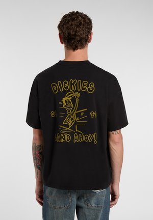 Man wearing black t-shirt with yellow cartoon character and text "Dickies Land Ahoy! 1922" on the back, paired with blue jeans.