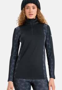 Junge Frau trägt ein schwarzes Half-Zip-Oberteil mit gemusterten Ärmeln und passenden Leggings, ergänzt durch eine schwarze Strickmütze.