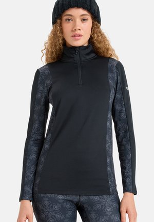 Young woman wearing a black half-zip top with patterned sleeves and matching leggings, accessorized with a black knit beanie.