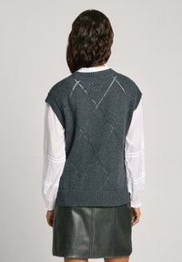 Gilet grigio lavorato a maglia con maniche corte, con un motivo a rombi e orlo a costine. Indossato sopra una camicia bianca a maniche lunghe, abbinato a una gonna verde scuro.