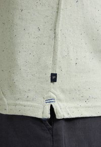 T-shirt in cotone verde chiaro con spruzzi di blu e nero. Presenta un orlo a spacco e un piccolo etichetta scura con un logo bianco.