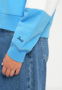 Persona con sudadera azul y blanca con "Levi's" bordado en el puño de la manga y jeans de mezclilla azul de cintura alta.