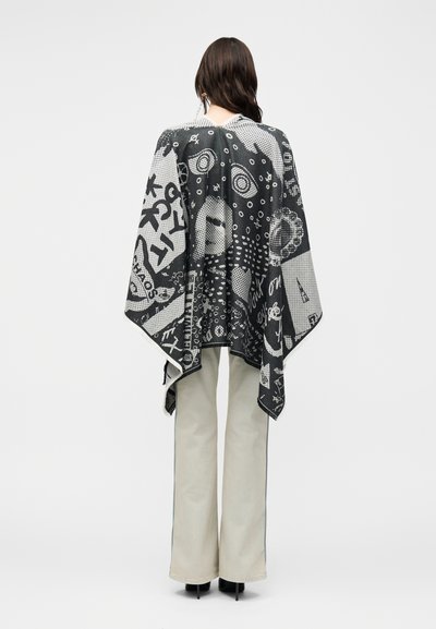 Γυναίκα που φοράει μαύρη και λευκή κάπα με αφηρημένα σχέδια, σε συνδυασμό με ανοιχτόχρωμο παντελόνι με φαρδιά μπατζάκια και μαύρα παπούτσια, με την πλάτη γυρισμένη.