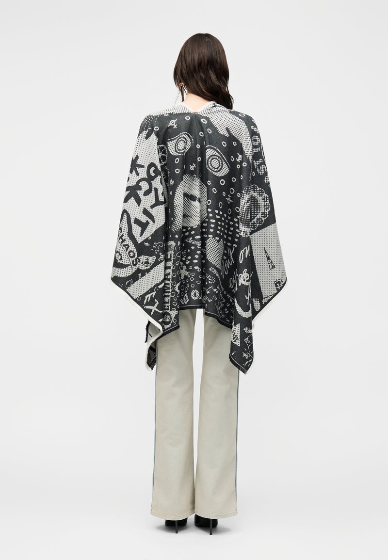 Femme portant une cape à motifs noir et blanc avec des dessins abstraits, associée à un pantalon large de couleur claire et des chaussures noires, vue de dos.