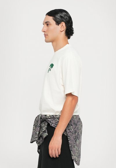 T-shirt branca de algodão com impressão gráfica verde, acompanhada de um tecido cinza com padrão atado na cintura. Corte reto, mangas curtas.