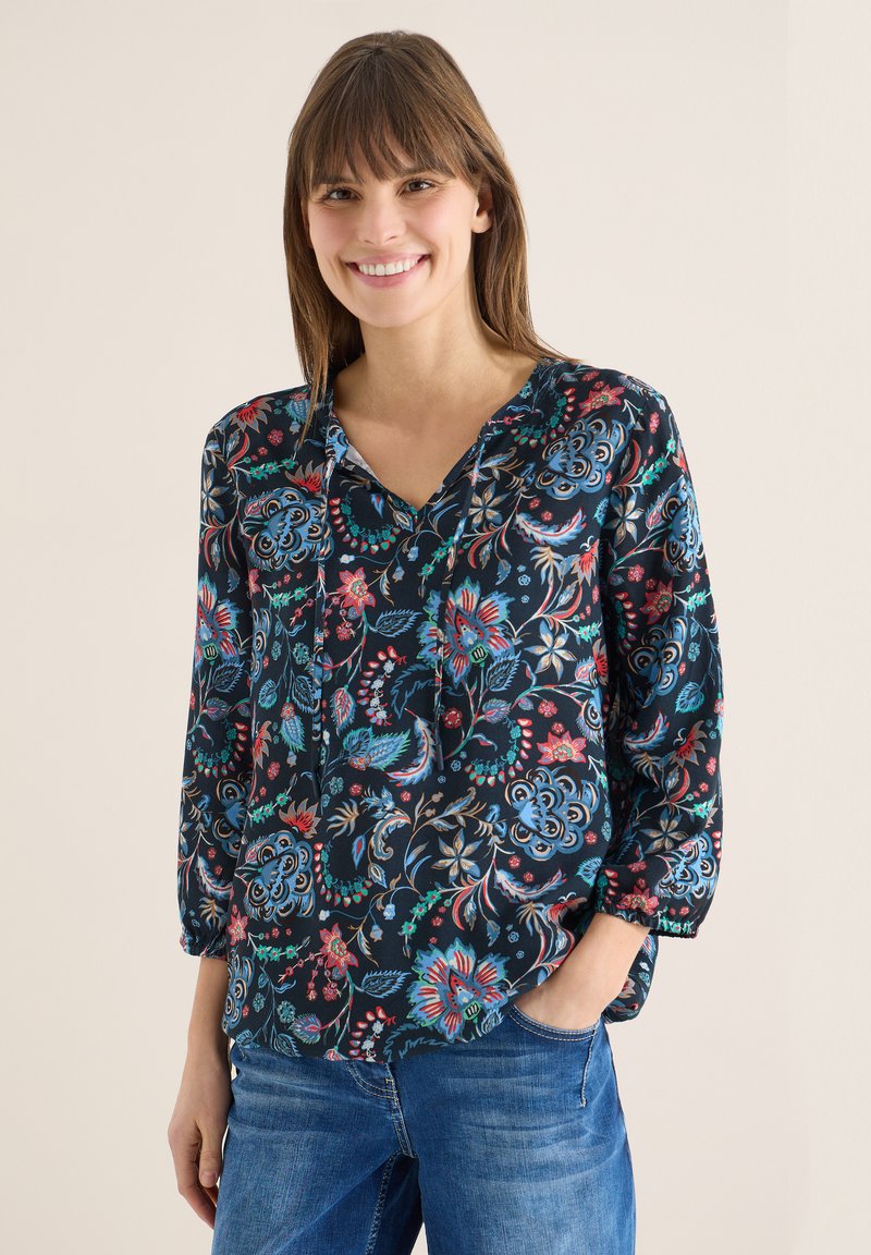 Femme souriante portant un chemisier à manches longues à fleurs foncées avec un col en V et un jean bleu, debout avec une main dans la poche.