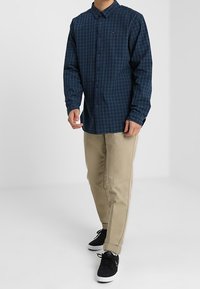 Blå och grön rutigt långärmad skjorta med knäppning fram, kombinerad med ljusbeiga byxor och svarta sneakers.