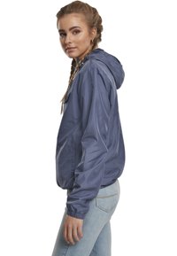 Giacca leggera con cappuccio di colore navy, con polsini elasticizzati e vestibilità rilassata. Dotata di due tasche laterali e una texture liscia e lucida.