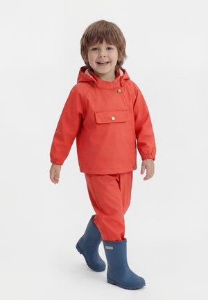 Young child smiling and walking, wearing an orange rain jacket, matching pants, and blue rubber rain boots on a white background.