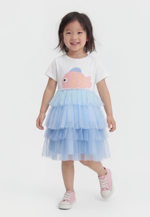 Χαμογελαστή νεαρή κοπέλα που περπατά, φορώντας λευκό πουκάμισο με ροζ σχέδιο ψαριού, στρωτή μπλε φούστα από τούλι και ροζ αθλητικά παπούτσια σε λευκό φόντο.