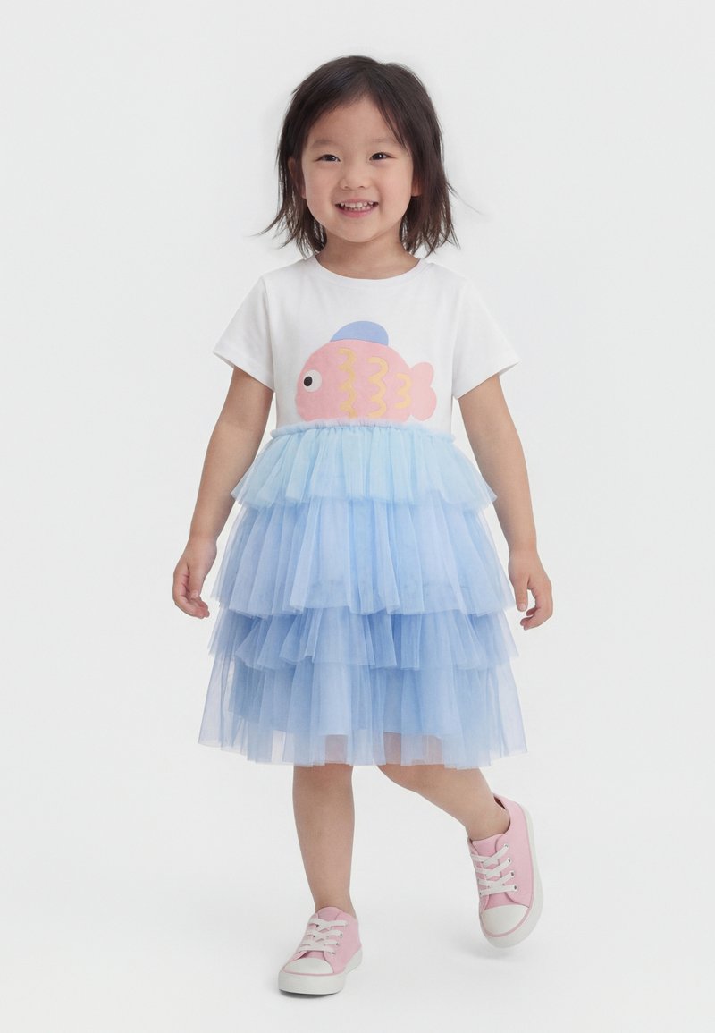 Χαμογελαστή νεαρή κοπέλα που περπατά, φορώντας λευκό πουκάμισο με ροζ σχέδιο ψαριού, στρωτή μπλε φούστα από τούλι και ροζ αθλητικά παπούτσια σε λευκό φόντο.