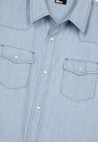 Camicia in denim blu chiaro con due tasche frontali, bottoni a pressione argentati e cuciture a contrasto. Collo classico e texture morbida.