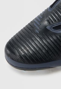 Μύτη μαύρου ποδοσφαιρικού παπουτσιού με υφή, που διαθέτει ραβδώσεις και μπλε λεπτομέρειες. Κατασκευασμένο από συνθετικό υλικό με λείο φινίρισμα.