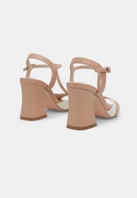 Sandales à talons beiges avec un talon bloc épais, un design découpé et des brides ajustables à la cheville. La tige est en similicuir lisse.