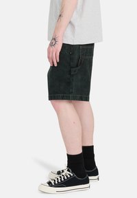 Personne portant un short en jean noir, des chaussettes noires, des baskets en toile noire avec des lacets blancs, et un T-shirt gris clair visible de profil.